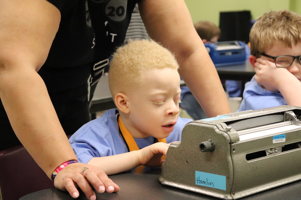 boy typing on a brailler with a teacher assisting him